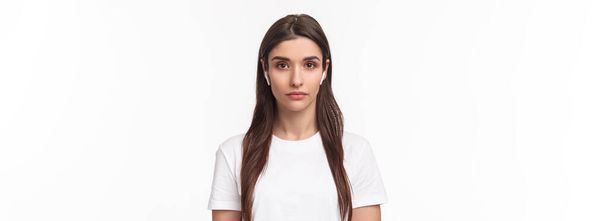 Zbliżenie portret poważnie wyglądającej atrakcyjnej białej kobiety w bezprzewodowych słuchawkach, patrzącej w kamerę z rozluźnioną twarzą bez emocji, stojącej w białym tle. Przestrzeń kopiowania - Zdjęcie, obraz