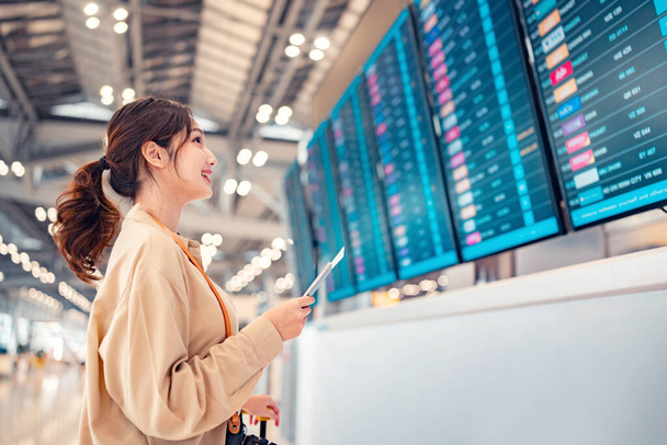 Felice viaggiatore donna asiatica controllando l'orario dei voli partenze bordo in aeroporto terminal hall di fronte al check-in banchi. Viaggio turistico concetto - Foto, immagini