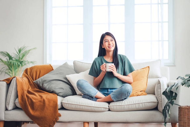 Happy young asian woman drinking coffee relaxing on sofa at home. Smiling female enjoying resting sitting on couch in modern living room. - Фото, изображение
