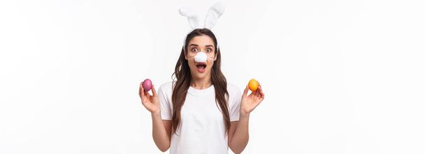 Пасхальная вечеринка начинается. Портрет смешной и милой, глупой молодой кавказской женщины, празднующей божественный день, носящей кроличьи уши и нос, держащей цветные яйца, задыхающейся, на белом фоне. - Фото, изображение