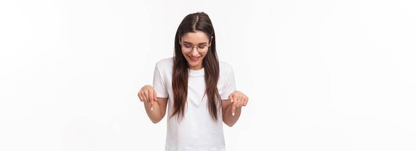 ウエストアップの肖像画の熱狂的な、笑顔幸せなブルネットの女性学生で眼鏡とTシャツ、下の広告を下を向いて見て、喜んで、白い背景を立って. - 写真・画像