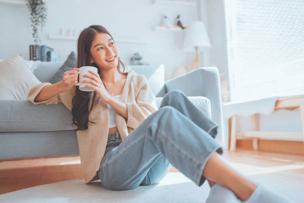 Happy young asian woman drinking coffee relaxing on the rug beside to the sofa at home. Smiling female enjoying resting sitting on couch in modern living room. - Photo, Image