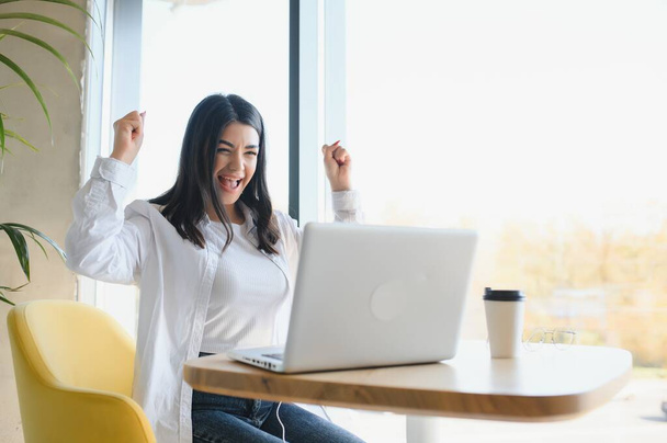 Girl rejoices in winning having raised her hands up sitting in front of laptop.Girl sitting in cafe, watches online sports match on computer.Woman blogger enjoys lot of views of her videos on Internet - 写真・画像