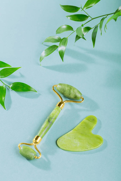 Πράσινο νεφρίτη ρολό προσώπου και gua sha ξύστρα με φύλλα σε γαλάζιο φόντο. Εργαλείο μασάζ για περιποίηση προσώπου, SPA αυτο-θεραπεία και περιποίηση ομορφιάς έννοια. - Φωτογραφία, εικόνα