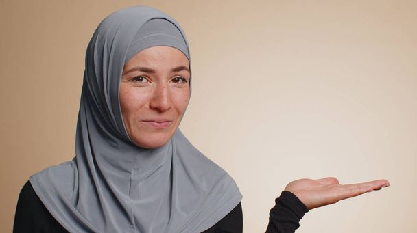 Mujer musulmana en hiyab mostrando pulgares hacia arriba y señalando lugar vacío, área de publicidad de texto comercial, espacio de copia para la promoción de bienes. Jovencita adulta. Estudio interior sobre fondo de pared beige - Foto, Imagen