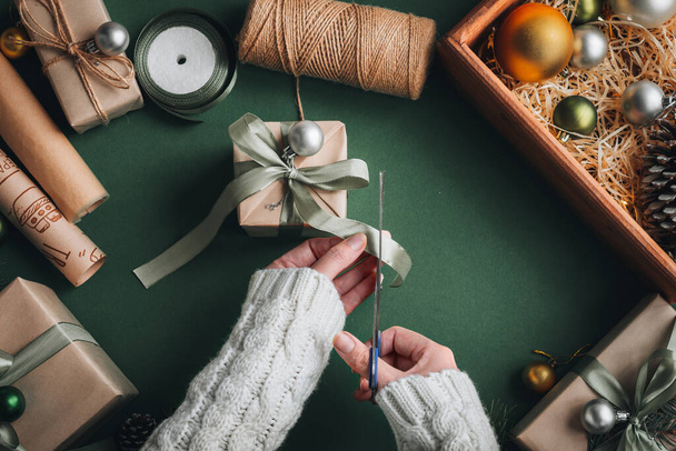 Creative hobby. Woman's hands wrap christmas holiday handmade present in craft paper with twine ribbon. Making bow at xmas gift box, decorated on olive table, top view. - Foto, imagen