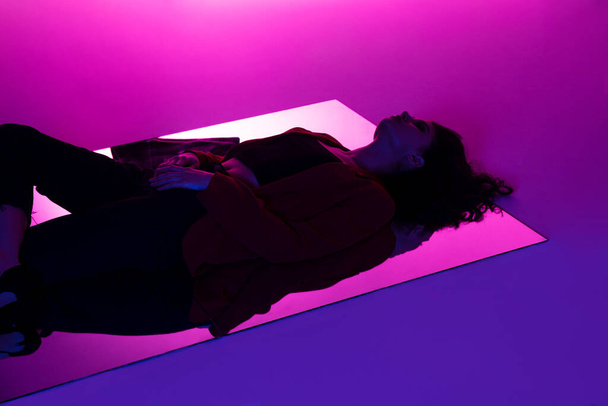 Sexy brunette young model in red suit and black jeans in mixed pink and violet neon light, lying down on mirror. Conceptual fashion photo. - Foto, Bild