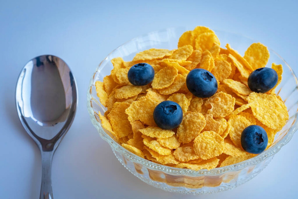delicioso muesli de maíz dulce y saludable con bayas de arándano en un tazón de cristal sobre un fondo azul - Foto, Imagen