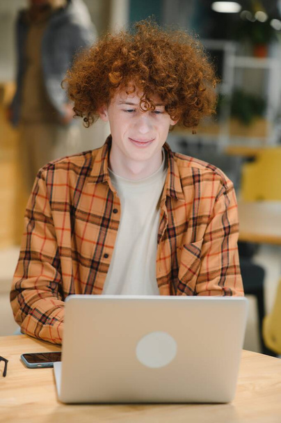 Young happy male freelancer in casual clothes with toothy smile looking at camera sitting in cafe with laptop and using mobile phone - Photo, Image