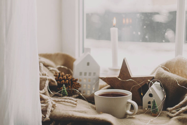 Чашка горячего чая, свечи, огни, маленький рождественский домик и елка, деревянная звезда на уютном одеяле на подоконнике. Зимний хигге. Атмосферный рождественский фон. Уютный дом в снежный день - Фото, изображение