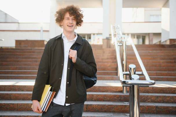 University.Smiling young student man holding a book and a bag on a university background .Young smiling student outdoors Life style.City.Student - Фото, зображення