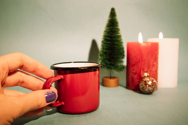 クリスマスツリーの背景に美しい紫色のマニキュアと女性の手の中に赤いマグカップコーヒーティーカップ、ろうそくを燃焼。2023年のお祝い。メリークリスマス、冬の休日は居心地の良い家でリラックス - 写真・画像