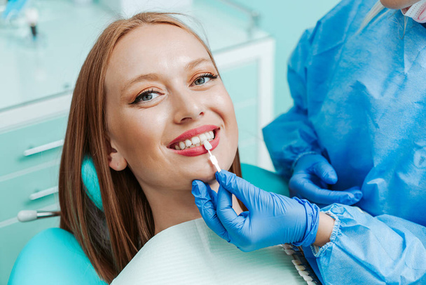Vonzó fiatal nő természetes fehér fogakkal a fogászati klinikán. Kezek orvos fogorvos fogak színpaletta mellett az arc. Mosoly egészséges fogak koncepció - Fotó, kép