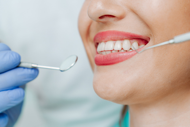 Dişçi kliniğinde açık mavi arka planda doğal beyaz dişleri olan gülümseyen bir kadın. Tıbbi aletlerle el doktoru diş hekimi. Sağlıklı diş konsepti - Fotoğraf, Görsel