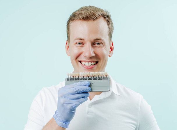 Nuori mies hymyilee luonnollisen valkoiset hampaat vaaleansininen tausta hammashoitola. Kädet pitäen hampaita väripaletti seuraavaksi kasvotusten. Hymyile terveet hampaat käsite - Valokuva, kuva