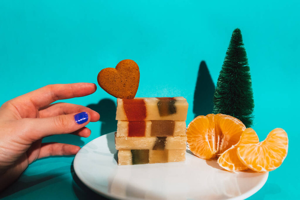 Spaans dessert tourron, turron, turrones op wit bord op tafel, vrouwenhand reikt naar marzepans. Nieuwjaar 2023 voedsel snoep, woestijn, mandarijn, hartvormige koekje blauwe achtergrond kerstboom  - Foto, afbeelding