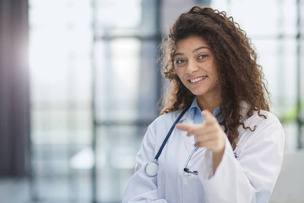 Uśmiechnięta młoda lekarka w białym płaszczu stojąca i wskazująca palcem do przodu - Zdjęcie, obraz