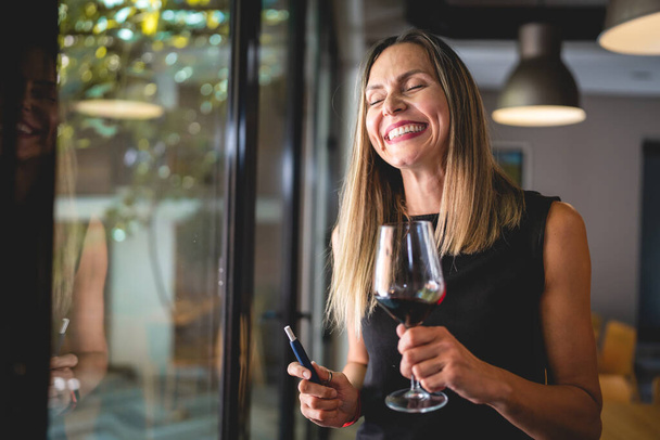 One woman adult middle age caucasian female standing by the window at home or restaurant alone holding glass of red wine and electronic cigarette device happy smile real people copy space - Photo, Image