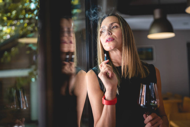 One woman adult middle age caucasian female standing by the window at home or restaurant alone holding glass of red wine and electronic cigarette device looking trough window real people copy space - Photo, Image