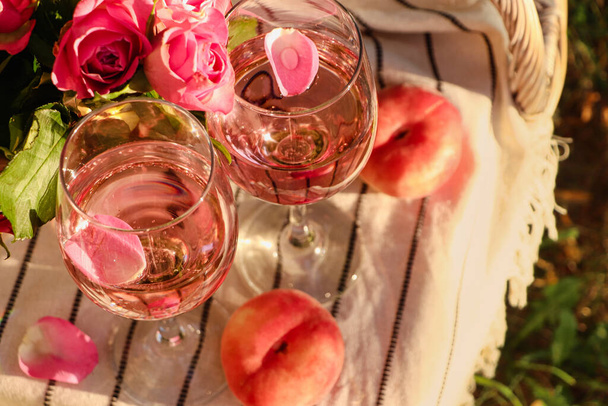 Glasses of delicious rose wine with petals, flowers and peaches on white picnic blanket outside, above view - Foto, Imagen