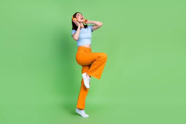 Full size photo of pretty positive girl stand one leg arms touch headphones isolated on green color background. - Zdjęcie, obraz