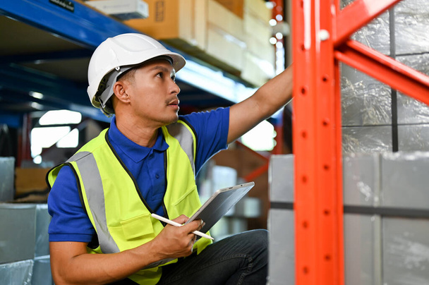 Концентрированный и профессиональный азиатский работник склада или бригадир в белой каске и униформе, проверяющий инвентарь, работающий на складе. - Фото, изображение