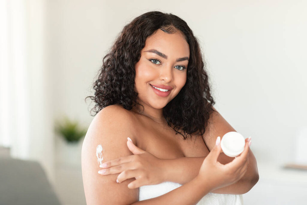 Fresh skin, moisturizing and nutrition for body. Positive african american plus size lady wrapped in towel using cream for skin in morning at home and smiling, empty space - Foto, afbeelding