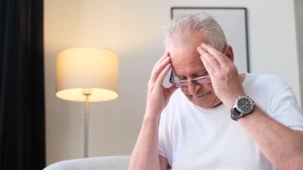Um idoso aposentado sentado no sofá em casa tem dor de cabeça. Depressão e fadiga. 4k vídeo - Filmagem, Vídeo