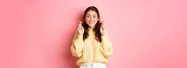 Image of cute cheerful woman cross her fingers for good luck and looking hopeful at camera, making wish, standing over pink background. - Fotó, kép