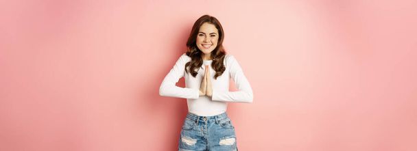 Cute modern girl with dark curly hairstyle, say please, thank you, holding hands in namaste thanks gesture, standing over pink background. - Foto, imagen