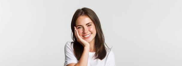 Close-up of beautiful smiling brunette girl touching cheeks, blushing over white background. - Фото, изображение