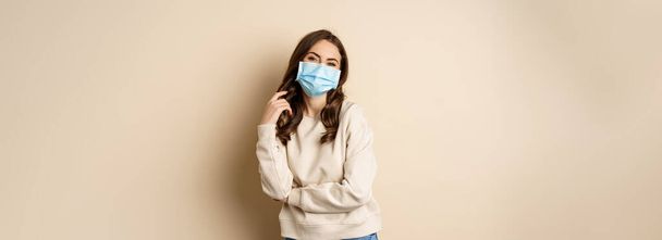 Covid-19, pandemic and quarantine concept. Beautiful modern woman in medical face mask, cross arms on chest, looking confident, beige background. - Foto, imagen