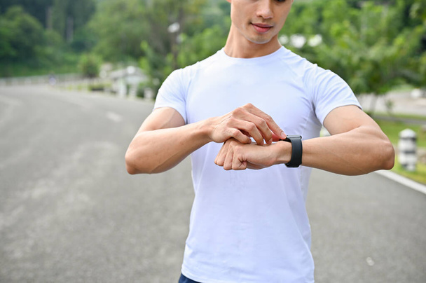 Красивый и атлетичный молодой азиат в спортивной одежде смотрит на свои умные часы, устанавливая программу подсчета калорий на свои спортивные часы. обрезанное изображение - Фото, изображение