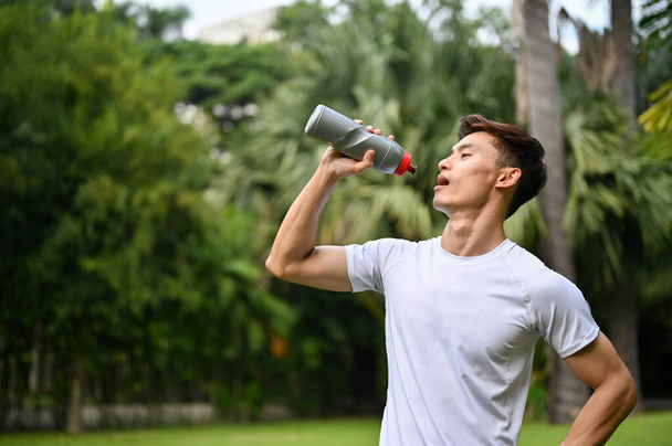 公園でのトレーニングの後、スポーツウェア飲料水やタンパク質飲料のハンサムな、喉の渇きと汗ばむ千年のアジアの運動選手. - 写真・画像