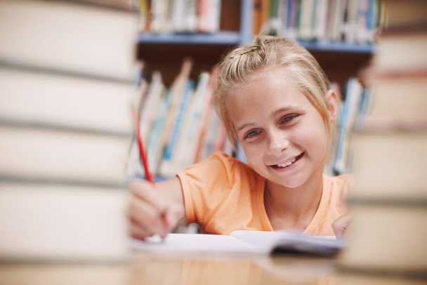 Le encantan sus estudios. Una joven feliz haciendo tareas escolares rodeada de libros en la biblioteca - Foto, Imagen