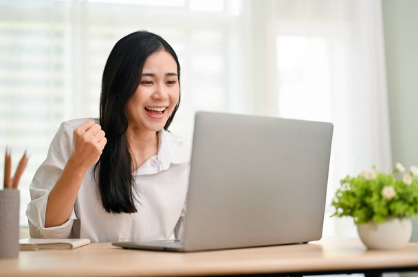 Успешная и жизнерадостная молодая азиатская студентка колледжа проходит онлайн-тест, получает предложение работы по электронной почте, празднует свой успех. Бизнесвумен, празднующая свой проект, одобрена за столом. - Фото, изображение
