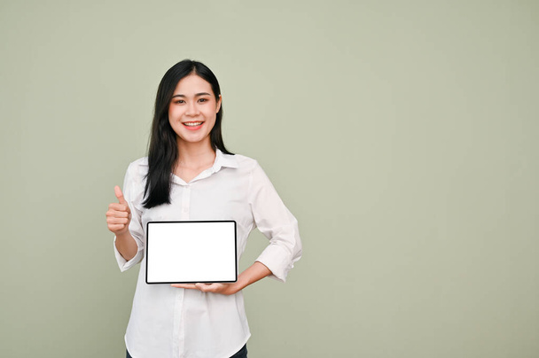 Beautiful young Asian woman in white shirt holding a digital tablet white screen mockup and showing thumb up, standing against grey background. recommend or suggest pose. - Photo, image