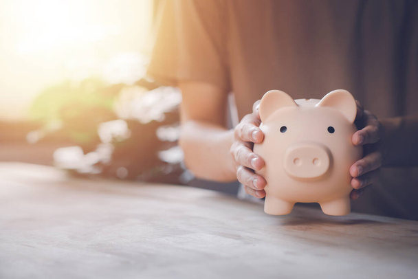 Man hand holding piggy bank on wood table. Saving money wealth and financial concept. Business, finance, investment, Financial planning. - 写真・画像