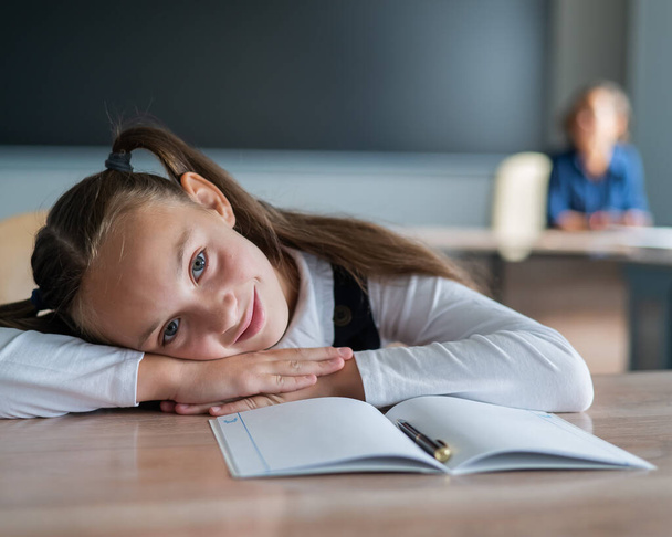 Pieni valkoihoinen tyttö on tylsistynyt koulussa. Koulutyttö taittoi päänsä pöydälle ja opettaja istuu taustalla - Valokuva, kuva