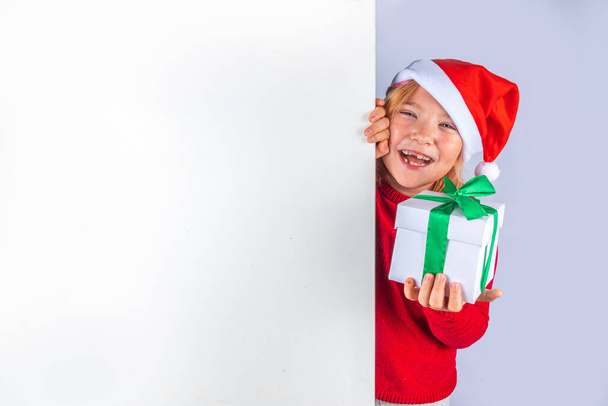 Christmas holiday background with funny blond girl. Little kid girl in red Christmas sweater and funny santa hat costume, with gift box on white background, Xmas greeting card, copy space - Fotoğraf, Görsel