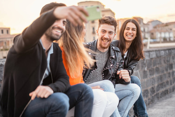 Group of young friends having fun sitting outdoors and taking memories with their smartphones at sunset - people and technology lifestyle concept - main focus on the brunette girl on the right - Foto, imagen