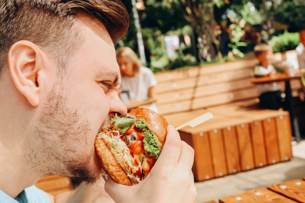 Giovane uomo caucasico affamato mangiare hamburger o hamburger di manzo con verdure e salsa in caffè all'aperto. Pranzo nella giornata di sole. Primo piano, fastfood, ritratto. - Foto, immagini