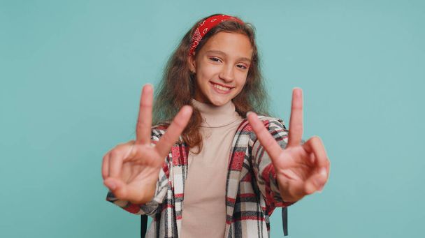 Happy teenager child girl kid showing victory sign, hoping for success and win, doing peace gesture smiling with kind optimistic expression. Joyful preteen children isolated on studio blue background - Photo, Image