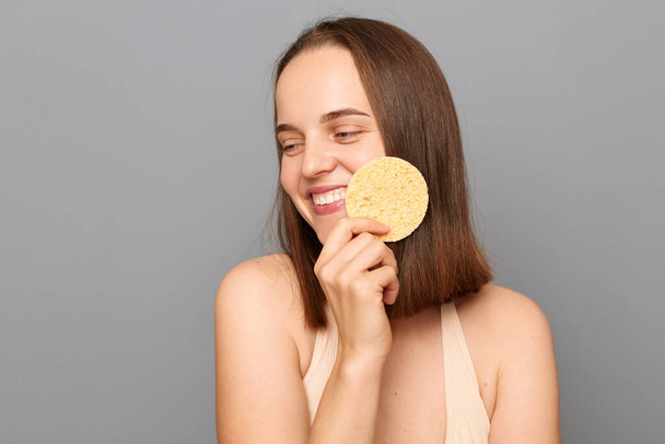 Photo of happy young adult girl with gentle smile holding cosmetic sponge near face isolated on gray background, looking away, enjoying cosmetic procedures. - Foto, Imagem