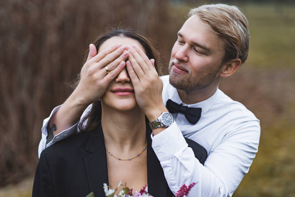 Bearded blond handsome Scandinavian groom in his late 20s covering the eyes of his beloved Middle Eastern wife. Medium closeup outdoor portrait. Love and romance. High quality photo - Φωτογραφία, εικόνα