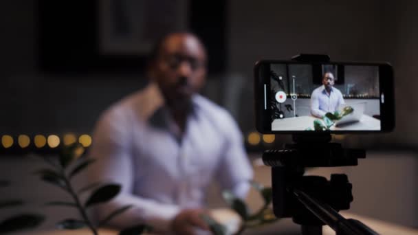 Afrikai férfi rögzíti video blog podcast segítségével okostelefon háromlábon beszélni távoli e-tanulás.Blogger befolyásoló, hogy online tanfolyam ül az asztalnál.Fekete ember teszi video leckét. Kiváló minőség - Felvétel, videó