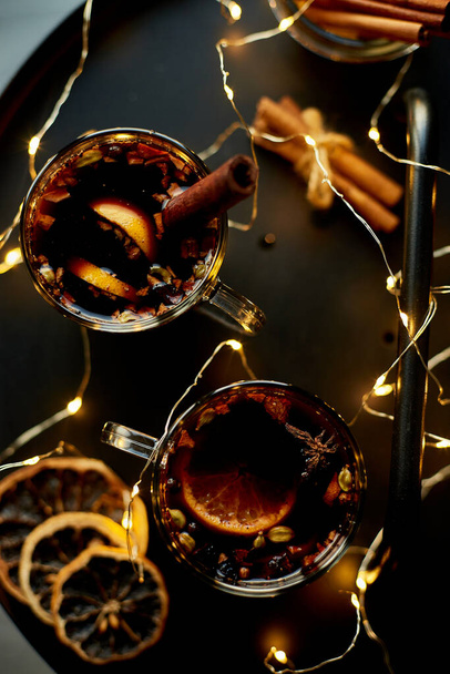 Świąteczne grzane czerwone wino z przyprawami i pomarańczami na czarnym stole z lekką girlandą, tradycyjny gorący napój na Boże Narodzenie, świąteczny koktajl - Zdjęcie, obraz