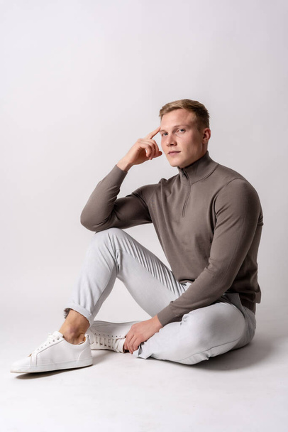 Portrait of an attractive blond Caucasian model in a brown sweater on a white background, sitting - Foto, Imagen