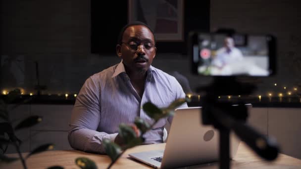 Africain homme enregistrement vidéo blog podcast à l'aide d'un smartphone sur trépied pour parler e-learning distant.influenceur blogueur faire cours en ligne assis à table.Black personne fait leçon vidéo. Haute qualité - Séquence, vidéo