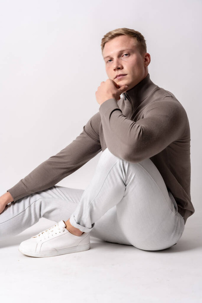 Caucasian blond model with a brown sweater on a white background, sitting on the floor with a serious look - Photo, image